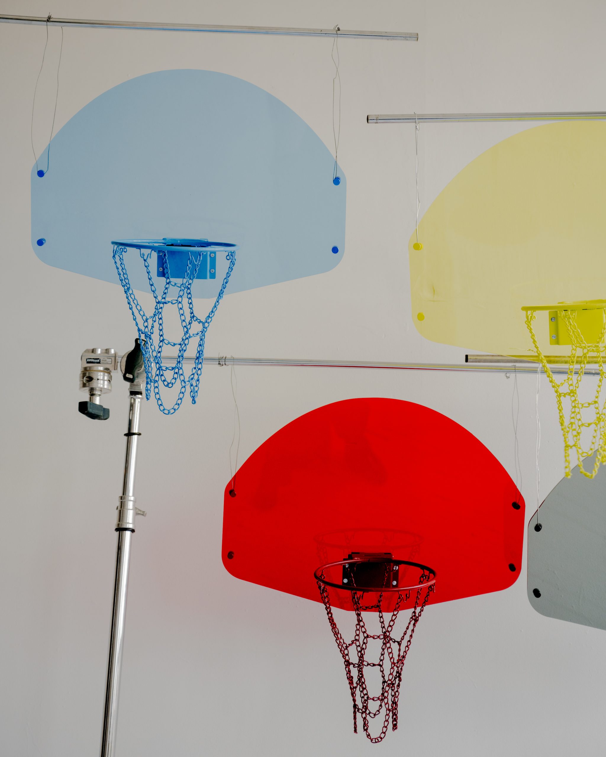 Three colorful transparent basketball backboards with chains as nets hang against a white wall; blue, yellow, and red backboards are shown. A metal stand supports one Acrylic Hoop - Blue (Copy).